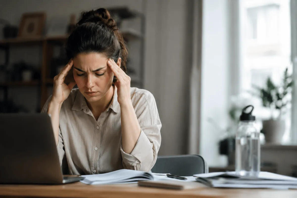 WFA-filtered water and cognitive function Woman sitting at a desk working on a laptop with documents spread out in front of her, holding her temples and appearing stressed or overwhelmed while reviewing paperwork in a home office setting.